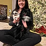 woman, dog, pomeranian, puppy, indoor, christmas_tree, holiday_lights, carpet, sitting, smiling, black_dog, white_dog, pet, home, decor, casual_clothing, leggings, shoes, wooden_sign, dog_bed