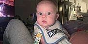 Hudson is registered to the contest to win money with this photo: baby, infant, face, bib, person, indoor, living_room, kitchen, television, furniture, refrigerator, couch, clothing, human, expression, curious, skin, head, eyes, resting