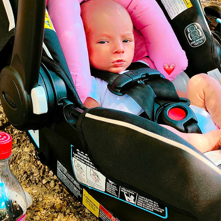 Nathaniel is registered to the contest to win money with this photo: baby, beverage, black_fabric, car_seat, child, face, ground, head_support, infant, outdoor, person, pink_cushion, plastic_bottle, resting, rocks, seatbelt, serious_expression, shadow, sunlight, warning_label