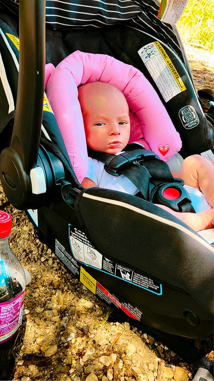 Nathaniel is registered to the contest to win money with this photo: baby, car_seat, pink_cushion, infant, seatbelt, outdoor, ground, rocks, plastic_bottle, beverage, sunlight, shadow, warning_label, black_fabric, child, person, face, head_support, resting, serious_expression
