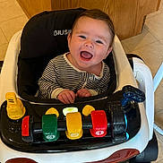 Preston is registered to the contest to win money with this photo: baby, baby_walker, toy, indoor, floor, wood_panel, tile_floor, smiling, child, striped_onesie, face, hands, happy, colorful, plastic, seat, walker_wheels, head, person, play