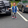 Princeton is registered to the contest to win money with this photo: toddler, child, running, laughing, parking_lot, car, denim_vest, sneakers, asphalt, outdoor, happy, joyful, casual_clothing, vehicle, person, road_marking, greenery, daylight, motion, smile