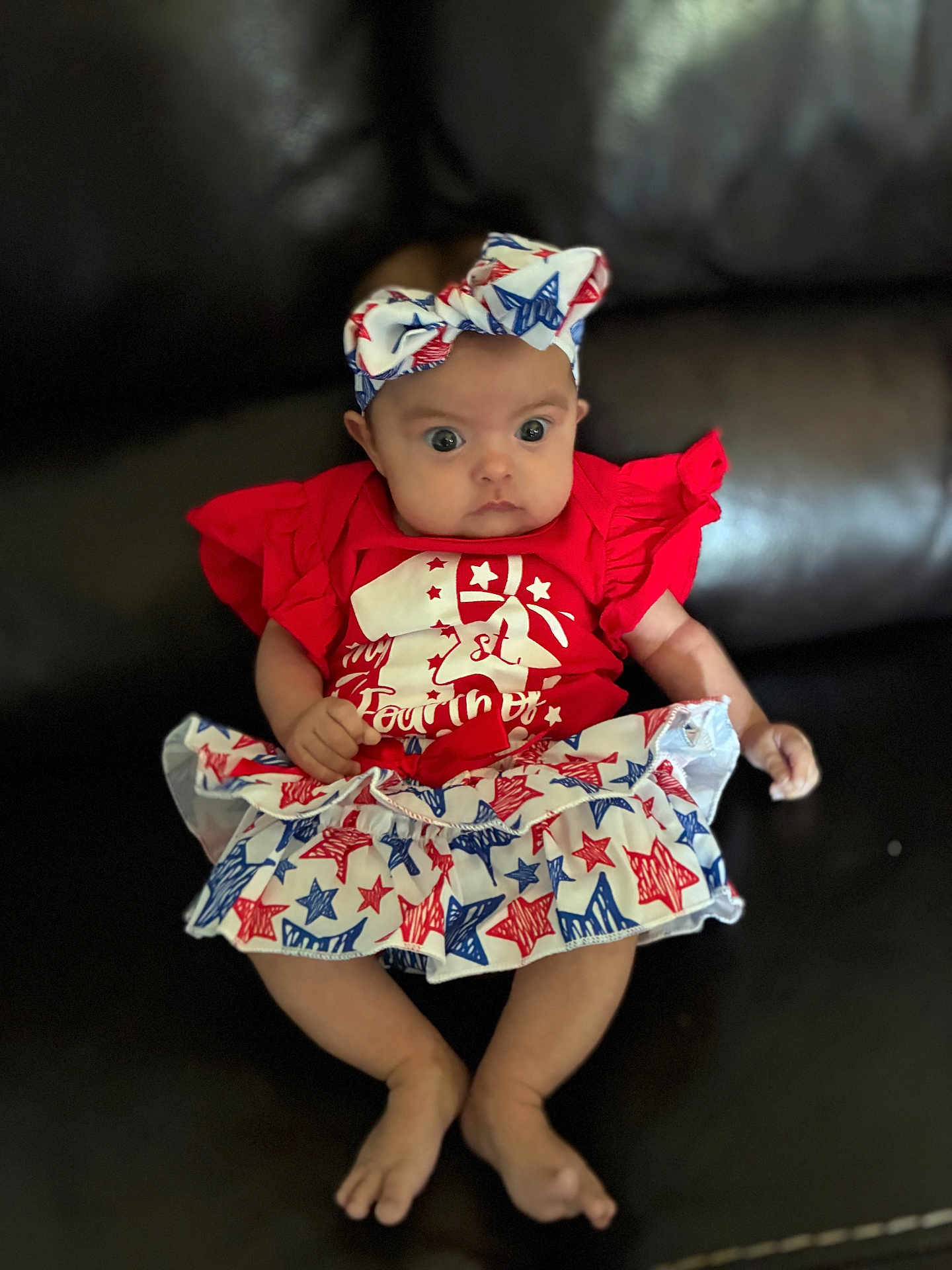Karmyn is registered to the contest to win money with this photo: baby, infant, child, red_clothing, headband, skirt, stars_pattern, sitting, leather_couch, curious_expression, wide_eyes, festive_outfit, indoor, person, cute, small_hands, barefoot, young, portrait, casual