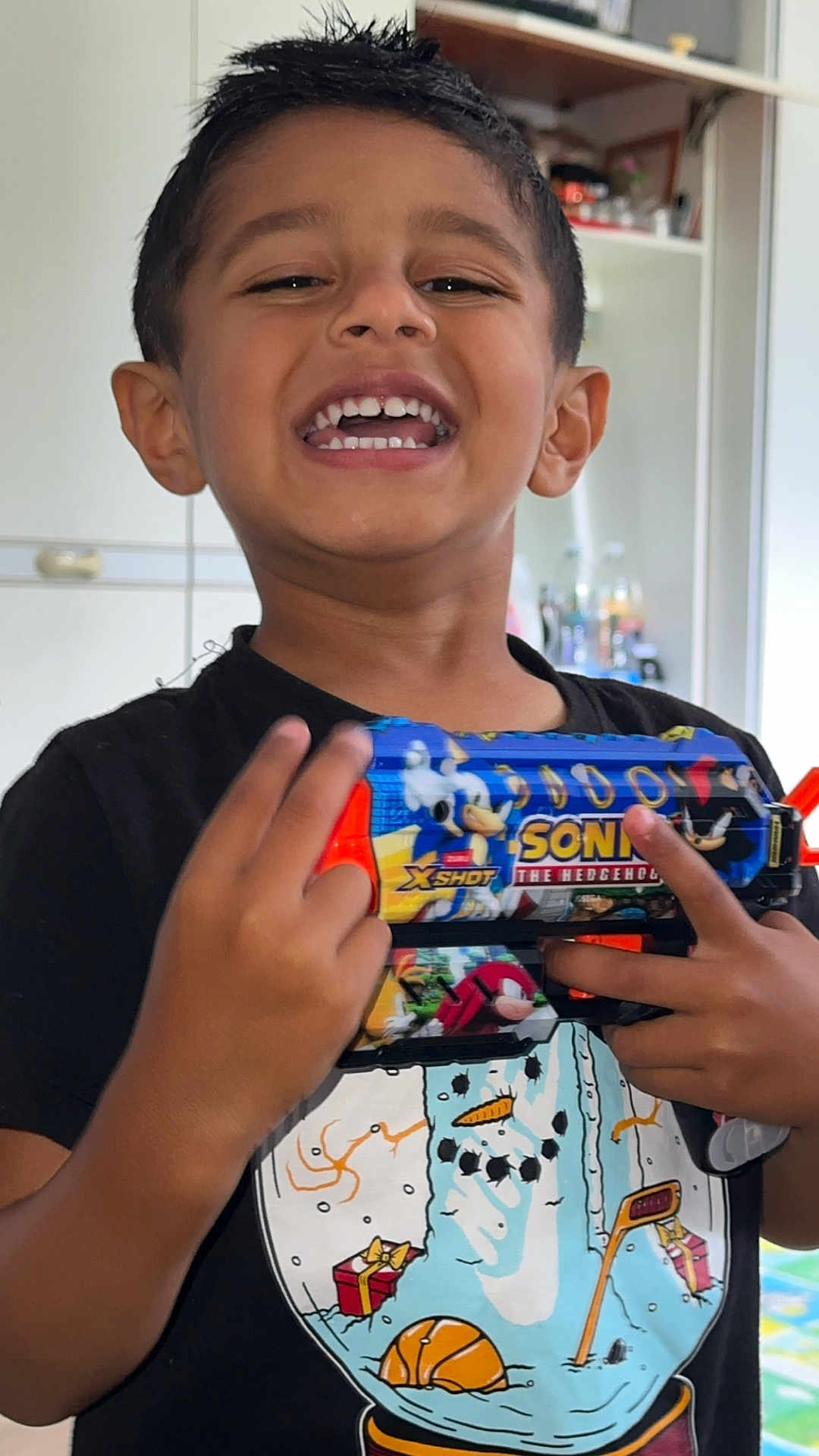 Zenil Patel joined the competition — help win amazing prizes! child, boy, smiling, toy_gun, sonic_the_hedgehog, indoor, black_tshirt, snowman_graphic, happy, portrait, person, short_hair, playful, hand, fingers, teeth, face, casual_clothing, background, home