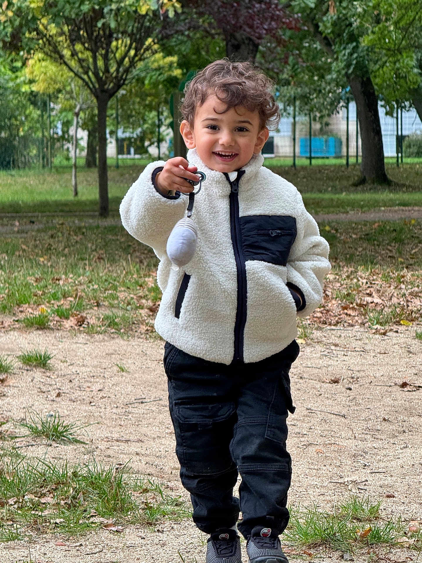 Isaac a rejoint le concours — aidez-le/la à gagner de superbes lots ! child, smiling, outdoor, park, curly_hair, jacket, pants, shoes, grass, trees, path, toy, keychain, happy, person, nature, casual_clothing, walking, daylight, playful
