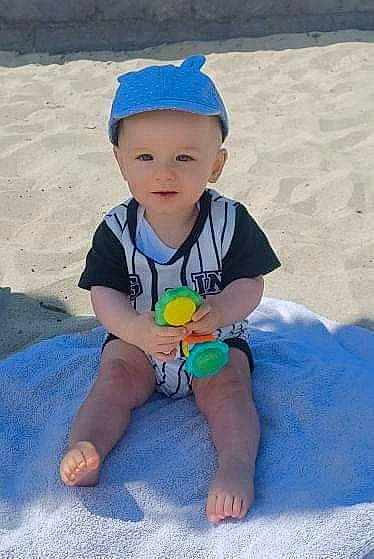 Brian participe au concours pour gagner de l'argent avec cette photo : baby, baby_toddler_clothing, barefoot, body_of_water, cap, electric_blue, eye, foot, fun, happy, head, headwear, human_leg, leisure, person, sand, shorts, sitting, sleeve, t_shirt