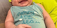 Stella is registered to the contest to win money with this photo: baby, smiling, infant, onesie, hand_gesture, couch, cushion, indoor, person, skin, face, expression, relaxed, young_child, text_on_clothing, blue_clothing, yellow_cushion, red_cushion, cheerful, portrait