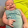 baby, smiling, infant, onesie, hand_gesture, couch, cushion, indoor, person, skin, face, expression, relaxed, young_child, text_on_clothing, blue_clothing, yellow_cushion, red_cushion, cheerful, portrait