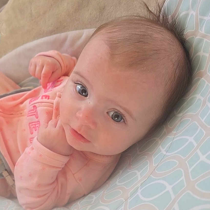 Elvira is registered to the contest to win money with this photo: baby, infant, face, eyes, hair, pink_clothing, cushion, pattern, lying_down, portrait, child, cute, soft_light, indoors, skin, hand, expression, young, cozy, closeup