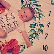 Elijah joined the competition — help win amazing prizes! baby, child, onesie, blanket, sloth, leaf_pattern, numbers, wreath, smiling, lying_down, infant, cute, humorous_text, playful, age_marker, indoors, soft_toys, portrait, skin, young_child