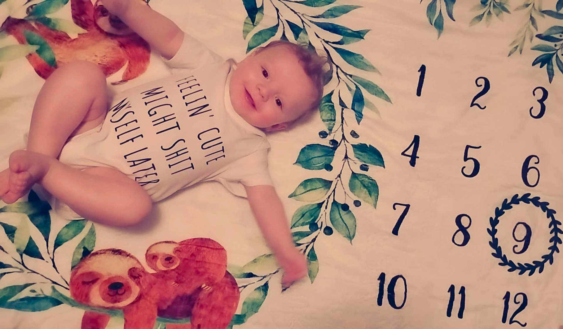 Elijah joined the competition — help win amazing prizes! baby, child, onesie, blanket, sloth, leaf_pattern, numbers, wreath, smiling, lying_down, infant, cute, humorous_text, playful, age_marker, indoors, soft_toys, portrait, skin, young_child