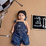 Ezekiel joined the competition — help win amazing prizes! baby, infant, child, portrait, lying_down, denim_overalls, beanie, black_hat, wrenches, tools, mechanic_theme, studio_photography, tan_background, letter_board, decorative_setup, wide_eyed, full_body, cute, fashion, milestone_photo