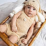baby, infant, child, portrait, face, wide_eyed, sitting, bassinet, wicker, knitwear, hat, romper, blanket, cozy, indoors, nursery, soft_lighting, brown, beige, photography