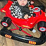 baby, child, toy_car, indoor, floor, red_couch, curly_hair, blue_eyes, clothing, black_and_white, checkered_pattern, plastic, wheels, seat, person, infant, wood_floor, furniture, playtime, expression