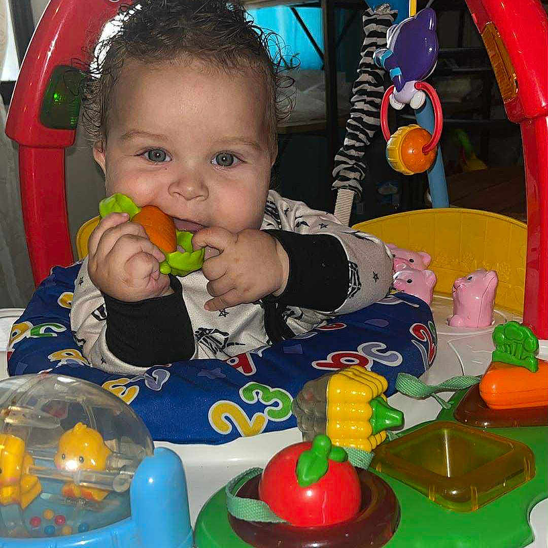 Malakai is registered to the contest to win money with this photo: activity_center, animal_toy, baby, blue_cushion, carrot_toy, child, colorful, curly_hair, face, green_toy, hands, indoor, infant, numbers, plastic_toy, play, red_frame, seat, smiling, toy