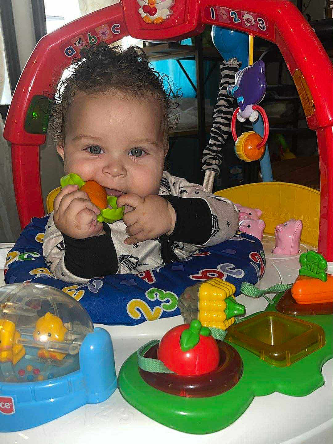 Malakai is registered to the contest to win money with this photo: baby, child, toy, activity_center, indoor, colorful, plastic_toy, carrot_toy, play, infant, curly_hair, smiling, face, hands, seat, numbers, animal_toy, red_frame, blue_cushion, green_toy