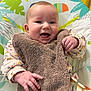 baby, smiling, infant, bib, baby_seat, toy, colorful, clothing, happy, face, hand, cute, indoor, child, playful, floral_pattern, soft_texture, person, young_child, expression