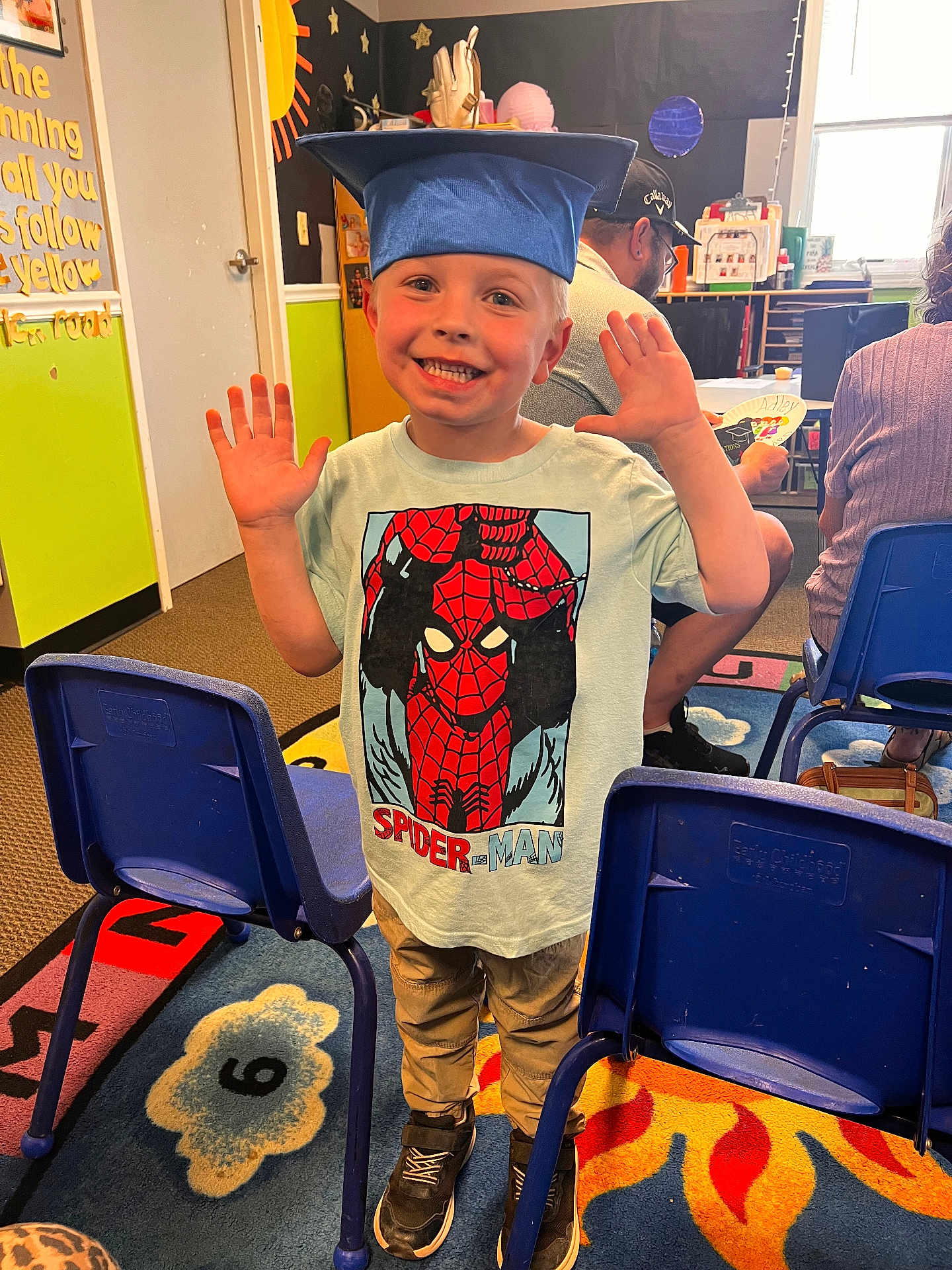 Carter is registered to the contest to win money with this photo: child, boy, graduation_cap, spiderman, tshirt, smiling, classroom, carpet, chairs, people, indoor, celebration, happy, learning, education, colorful, toys, furniture, person, casual_clothing