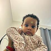 Summer is registered to the contest to win money with this photo: baby, child, curly_hair, floral_clothing, indoor, person, portrait, cute, looking_at_camera, hand, wristband, tattoo, curtains, wall, ceiling, soft_light, expression, young_child, holding, closeup
