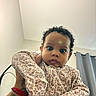 baby, child, curly_hair, floral_clothing, indoor, person, portrait, cute, looking_at_camera, hand, wristband, tattoo, curtains, wall, ceiling, soft_light, expression, young_child, holding, closeup