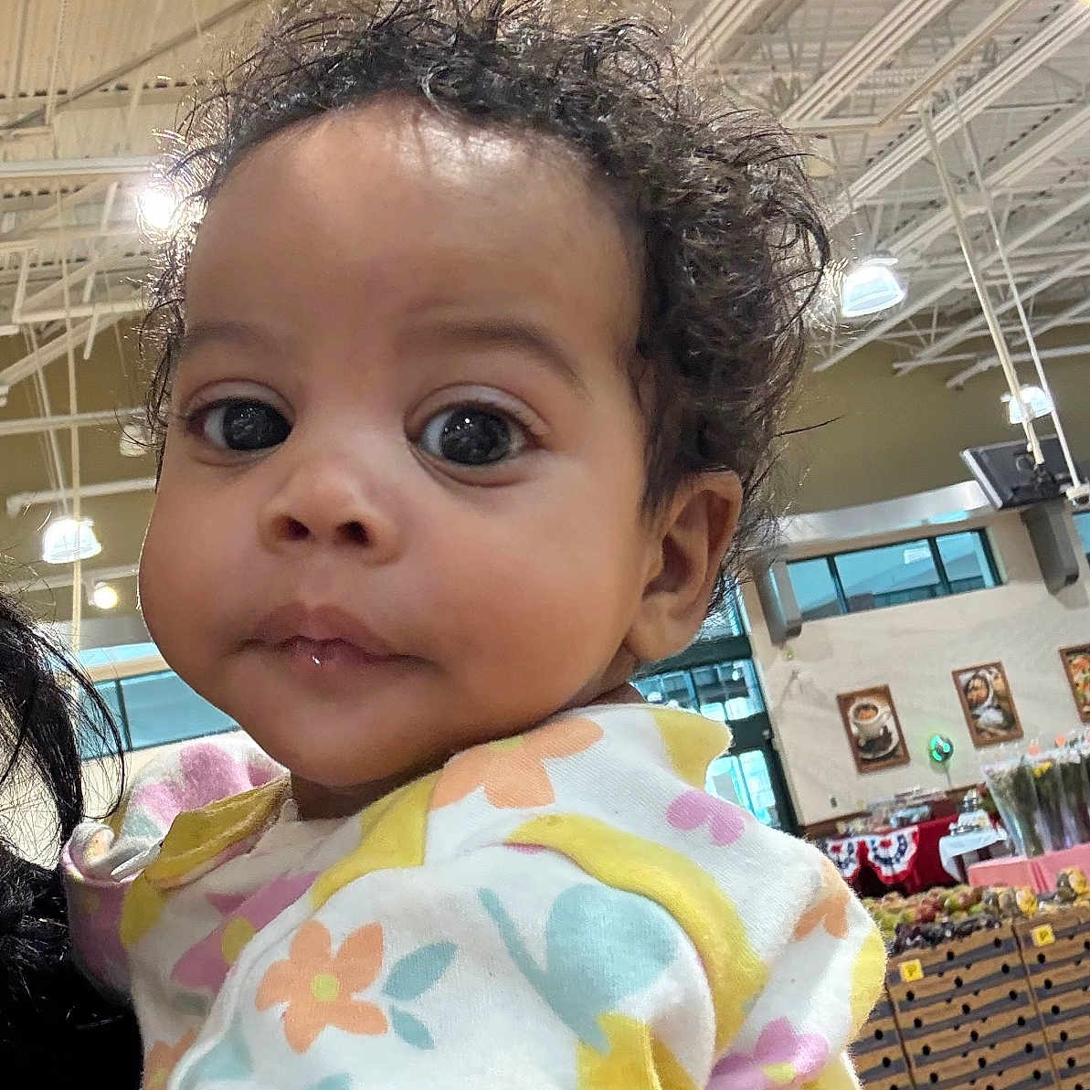 Summer is registered to the contest to win money with this photo: baby, child, curly_hair, indoor, market, grocery, colorful_clothing, floral_pattern, person, face, large_eyes, cute, holding, portrait, infant, bright_lighting, ceiling_lights, wooden_crates, shopping_area, curious_expression