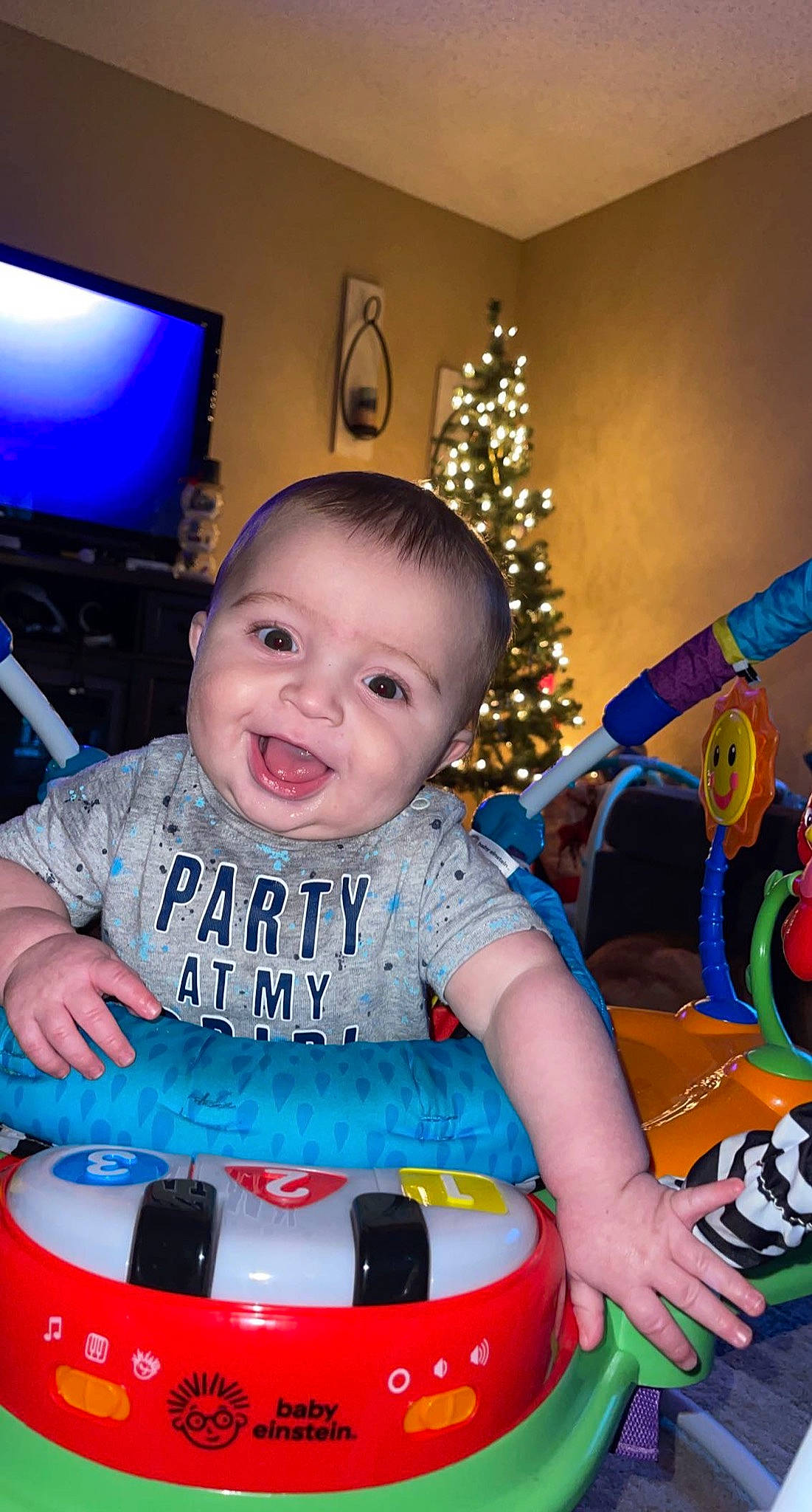 Kane is registered to the contest to win money with this photo: baby, baby_toddler_clothing, bumper, child, christmas_tree, event, eye, fun, happy, holiday, lap, leisure, lighting, person, recreation, riding_toy, sitting, smile, t_shirt, television