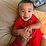 baby, child, infant, smiling, happy, portrait, red_outfit, bed, bedding, pillow, blanket, bedroom, indoor, adult_hand, family, sitting, close_up, dark_hair, brown_eyes, cozy