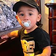 Elias joined the competition — help win amazing prizes! child, toddler, baby, pacifier, cap, black_shirt, sitting, indoor, portrait, face, eyes, short_hair, living_room, couch, cushion, wooden_furniture, home, warm_lighting, looking_at_camera, cute