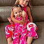 child, children, siblings, smiling, pink_clothing, couch, leather, happy, family, indoor, portrait, barefoot, toddler, cute, affection, embrace, young, hairclip, playful, seated