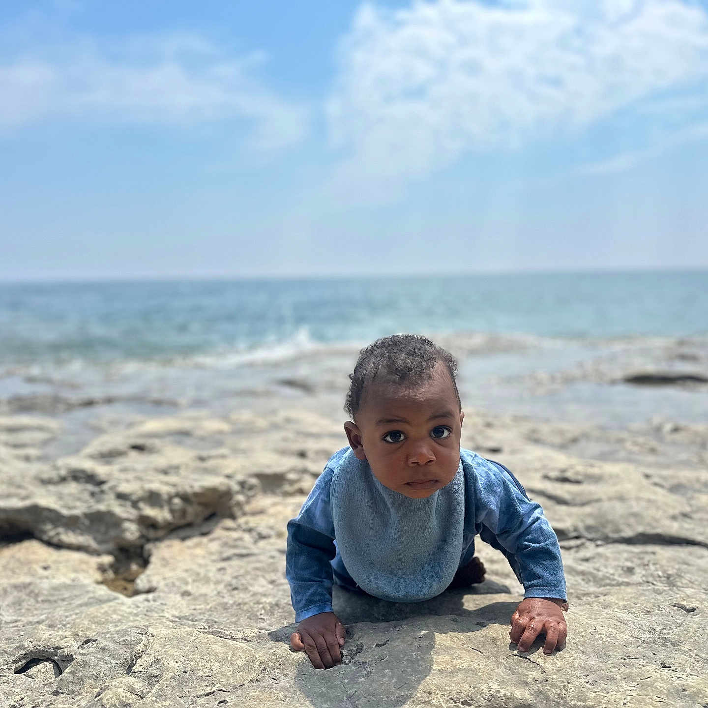 Xayvion joined the competition — help win amazing prizes! baby, blue_bib, child, clouds, crawling, curious, daylight, exploration, front_view, infant, nature, ocean, outdoor, person, portrait, rocky_surface, sea, sky, water, young_child