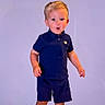 child, toddler, boy, standing, barefoot, portrait, fashion, outfit, navy_shirt, shorts, blond_hair, smiling, surprised, indoors, studio, lavender_background, full_body, person, photography, clothing