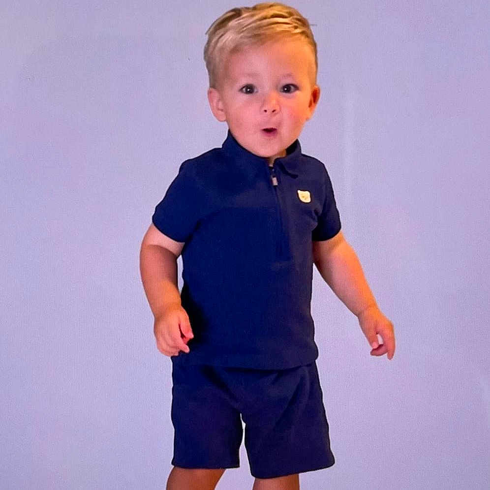 Charles is registered to the contest to win money with this photo: child, toddler, boy, standing, barefoot, portrait, fashion, outfit, navy_shirt, shorts, blond_hair, smiling, surprised, indoors, studio, lavender_background, full_body, person, photography, clothing