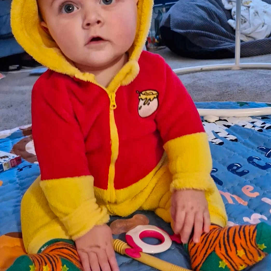 Albert is registered to the contest to win money with this photo: baby, carpet, child, curious, cute, expression, face, floor, hand, hooded_clothing, indoor, infant, person, play_mat, red, sitting, sock, striped_sock, toy, yellow