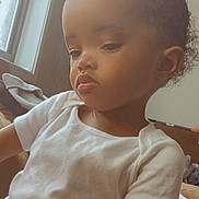 Nyomi is registered to the contest to win money with this photo: child, toddler, indoor, window, natural_light, white_shirt, peach_shorts, curly_hair, bun, serious_expression, sitting, couch, blanket, skin, face, person, portrait, young_child, home, casual_clothing