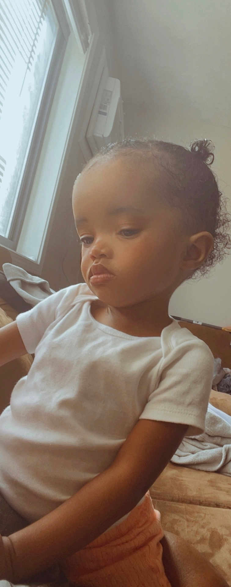 Nyomi is registered to the contest to win money with this photo: child, toddler, indoor, window, natural_light, white_shirt, peach_shorts, curly_hair, bun, serious_expression, sitting, couch, blanket, skin, face, person, portrait, young_child, home, casual_clothing