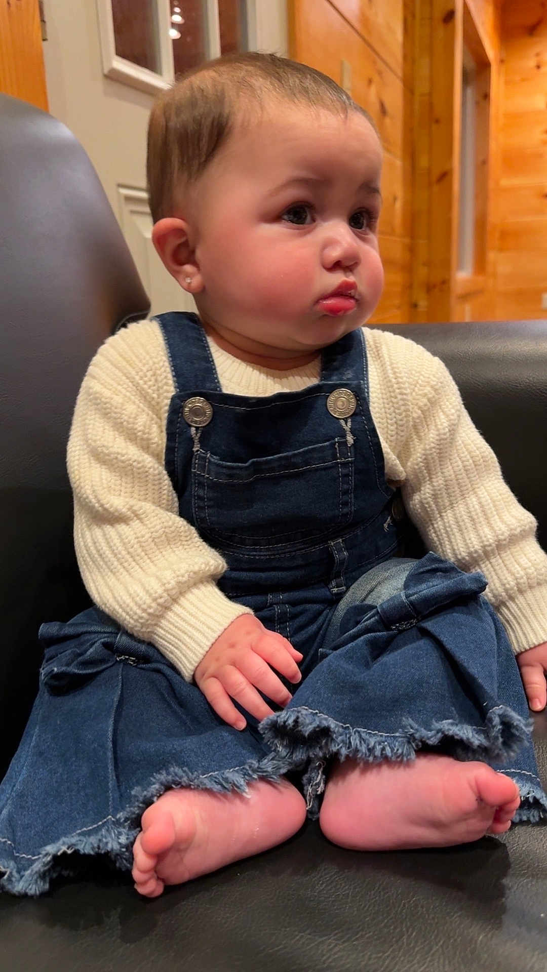 Teagan is registered to the contest to win money with this photo: baby, child, barefoot, denim, overalls, sweater, knitwear, indoor, furniture, chair, wood_paneling, cute, portrait, face, hands, feet, earrings, expression, seated, cozy