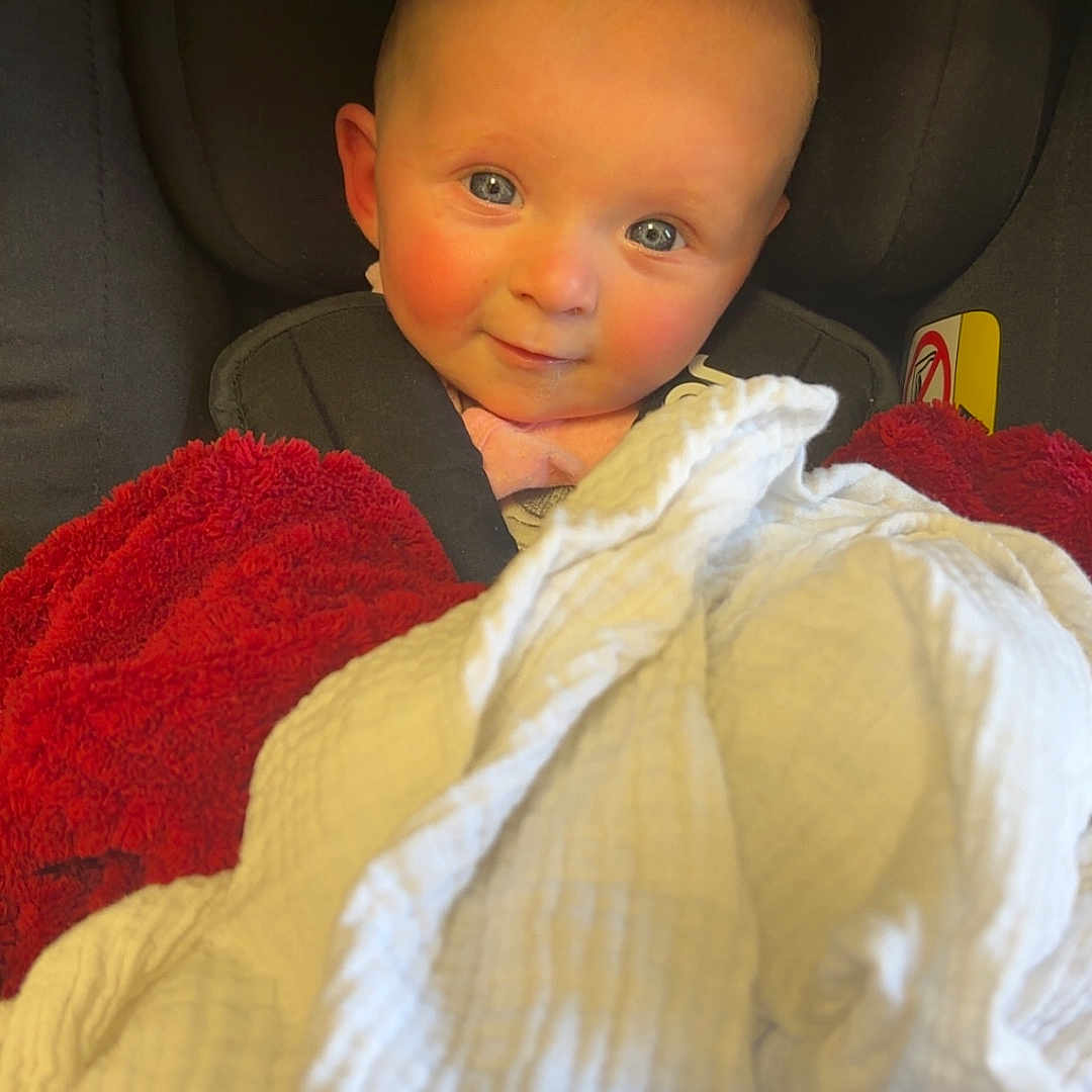Mercedes is registered to the contest to win money with this photo: baby, blanket, blue_eyes, car_seat, child, clothing, cozy, cute, face, head, indoor, infant, person, portrait, red_blanket, rosy_cheeks, seated, smile, white_blanket, young_child