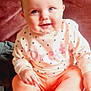baby, infant, smiling, blue_eyes, couch, pink_clothing, heart_pattern, sitting, indoors, cozy, cute, child, soft_lighting, portrait, plush_furniture, happy, person, young_child, warm_colors, casual
