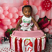Sariyah is registered to the contest to win money with this photo: baby, child, high_chair, crown, strawberry, balloon, pink, red, white, decorations, party, celebration, milestone, infant, smiling, face, clothing, floor, background, tassels