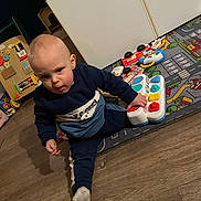 Eljy a rejoint le concours — aidez-le/la à gagner de superbes lots ! toddler, child, toy, floor, playmat, indoor, clothing, piano_toy, furniture, cabinet, wooden_floor, person, curious, sitting, sock, play, colorful, room, young_child, expression