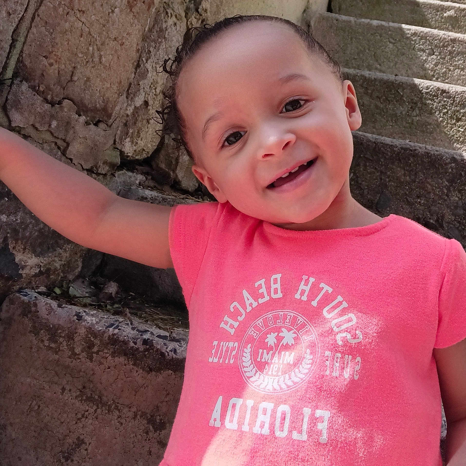 Ce'Ona joined the competition — help win amazing prizes! background, candid, casual_clothing, child, curly_hair, face, happy, joyful, natural_light, outdoor, person, pink_tshirt, portrait, rustic, smiling, stone_stairs, sunlight, texture, toddler, young_child