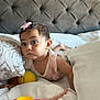 baby, child, toy, bed, blanket, pillow, headboard, pink_bow, curious, indoors, soft_light, infant, clothing, plaything, sleeping_area, toddler, cute, portrait, person, resting