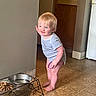Gunner is registered to the contest to win money with this photo: barefoot, blonde_hair, cabinet, child, curious_expression, dog_food, dog_water_bowl, holding_object, home, indoor, kitchen, metal_bowl_stand, person, playing, purple_ring, short_sleeve_onesie, standing, tile_floor, toddler, wall