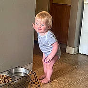 Gunner is registered to the contest to win money with this photo: barefoot, blonde_hair, blue_eyes, child, corner, curious, food_bowl, holding_object, home, indoor, kitchen, light, mischievous, onesie, pet_bowl, short_sleeve, standing, tile_floor, toddler, water_bowl