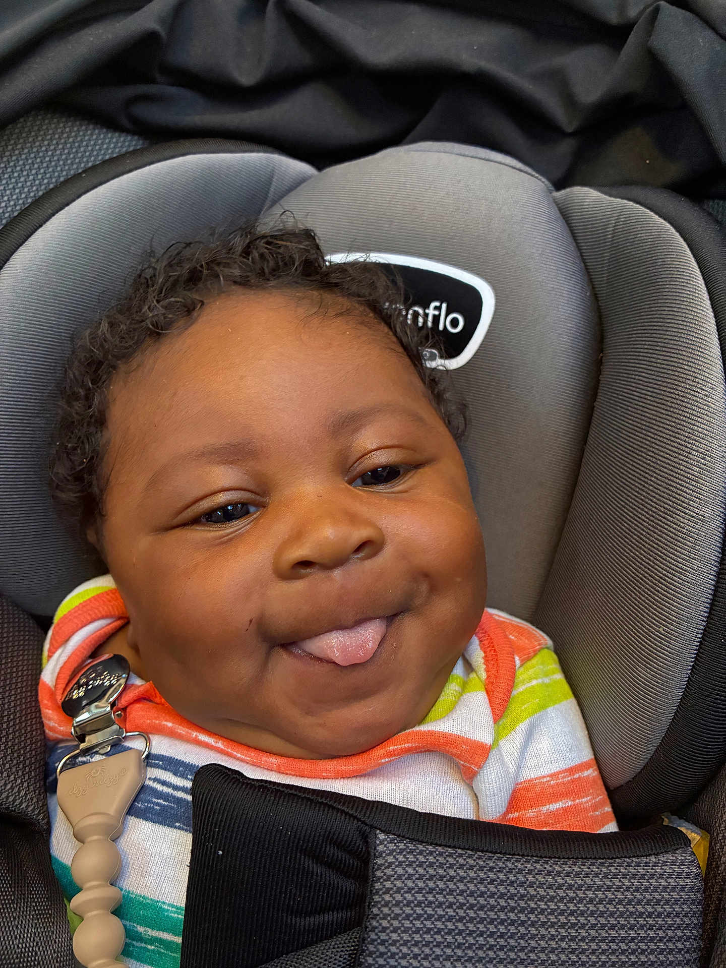 Kemet Brooks is registered to the contest to win money with this photo: baby, child, car_seat, tongue_out, smile, curly_hair, colorful_clothing, striped_shirt, black_blanket, cute, playful, infant, seatbelt, comfort, closeup, face, happy, indoors, person, portrait