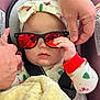 baby, infant, sunglasses, red_lenses, headband, bow, car_seat, seatbelt, pacifier, blanket, adult_hand, hand, reflection, holiday_pjs, christmas_tree_pattern, cozy, portrait, closeup, chubby_cheeks, accessory