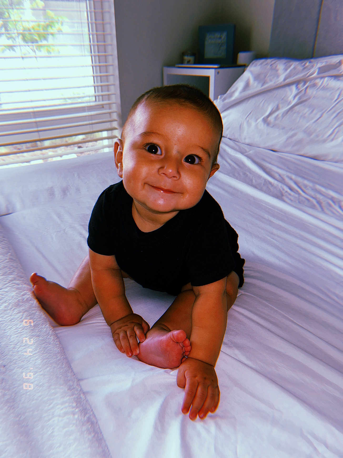Luka joined the competition — help win amazing prizes! baby, child, infant, smile, black_clothing, bed, white_bedspread, indoor, window, blinds, cabinet, foot, hand, curious_look, cheeky_smile, sitting, portrait, room, cozy, young_kid
