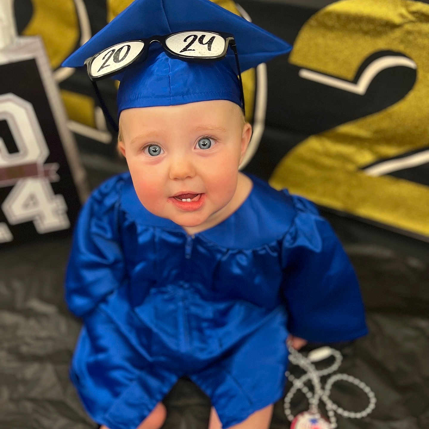 Innocence joined the competition — help win amazing prizes! baby, beaded_necklace, black_background, blue_clothing, celebration, child, cute, event, eyes, face, glasses, gold_numbers, graduation_cap, graduation_gown, indoor, medallion, portrait, sitting, smiling, white_numbers