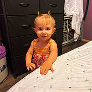 Anne is registered to the contest to win money with this photo: toddler, child, baby, smiling, portrait, indoors, bedroom, dress, hair_buns, standing, face, person, bed, dresser, drawers, laundry, baby_gate, home, furniture, photography