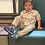 child, boy, indoor, bench, window, curtains, casual_clothing, palm_tree_pattern, sneakers, blue_shoes, beige_pants, teal_cushion, wood_floor, light, smiling, hair, face, relaxed, seated, daylight